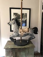 Full side view of gray carousel horse with black mane mounted on metal pole and black round base, placed on wooden cabinet in front of wall art.