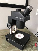 Black Bausch and Lomb compound microscope on greenish wood table, showing angled dual eyepieces, focus knob, and illuminated base with some wear.