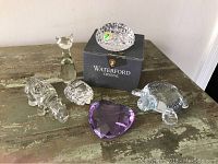 Overview of six glass and crystal items on wooden surface including Waterford crystal dish in box, purple heart paperweight, and various animal figurines