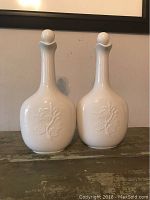 Pair of white vases/jugs with embossed floral pattern on bulbous bases, narrow necks with flared rims, and round stoppers on top.
