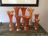 Front and angled view of seven Fenton carnival glass vases in shades of orange and amber with iridescent sheen on a rustic wooden table below a framed picture.