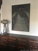 Framed oil painting hanging on wall above wooden cabinet. Shows the full artwork and surrounding area for context.