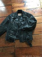 Front view of vintage black leather motorcycle jacket laid flat on wooden floor, showing front zipper closed with belt, quilted lining visible from collar, zippered pockets and zippered cuffs.