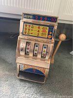 Front angled view of vintage toy slot machine with lever on side showing the spinning reels, payout info, coin slot and payout tray