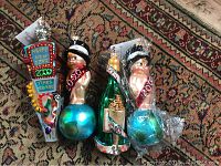 Four Christopher Radko New Year 2000 ornaments arranged on a carpet, including two baby figurines on blue globes, a Times Square sign, and a green champagne bottle, with one baby ornament wrapped in plastic.