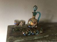 Front view of the vintage handpainted Italian art glass decanter with two wine glasses and three small handled glasses on a wooden surface.