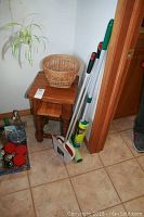 Overall view of small wooden table with woven basket on top, broom with dustpan and cleaning supplies nearby.