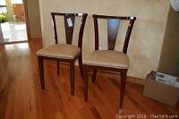 Two mid-century modern dining chairs with dark wood frames and beige upholstered seats placed on a wood floor against a light textured wall.