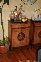 Front view of the antique look wooden cabinet with floral decoration on drawer and doors, black marble top, placed next to a potted plant and holding multiple decorative items on top.