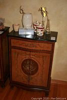 Antique look wooden cabinet with black marble top, floral decorations, drawer and doors, with decor arranged on the top.