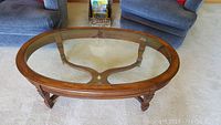 Oval solid walnut coffee table with clear glass top, sitting on beige carpet between two blue upholstered chairs.