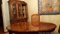 Photo showing polished oval solid fruitwood dining table with one chair and china cabinet in the background.