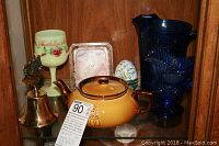 Photo showing ceramic teapot, brass bell, decorative wineglass, egg trinket box, and blue glass pitchers on glass shelf.