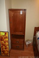 Tall Stanley wood cabinet with two drawers and a door, stained wood, standing against wall on wooden floor.