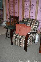 Photo of easy chair with wood arms and two orange throw pillows on seat and one gray pillow on side, with metal swivel bar stool and green cushioned seat next to it.