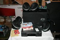 Full set view showing six Mirage Omni speakers, one subwoofer, Zenith VCR, remote, VHS tapes, and Monster power strip on white tablecloth.