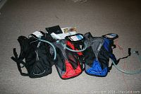 Three JanSport Helium hydration packs laid flat on carpet in black, red, and blue versions, showing adjustable straps and drinking tubes.