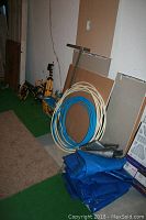 Photo of coiled white and blue tubing, folded blue tarps, leaning tile sheets and various small tools in a corner.