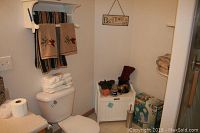 Full view of bathroom essentials: folded towels on toilet tank and shelf, small decorative plants and items on white cabinet, toilet paper rolls on counter, and a sign on wall.