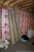 Three rolled berber carpets standing upright in an unfinished room with exposed insulation and framing.
