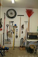 Wide shot of assorted outdoor tools hanging on pegboard including shovels, snow shovel, rakes, tree pruner, axe, pitchfork, and post pounder with some tools resting on floor and table on right side