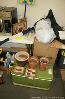 Photo showing grouped items including cooler, terra cotta pots, wrapped fabric bundle, garden plaques, and household items on table