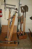 Full view of garden tools including various rakes, hoes, and shovels in a garage area.