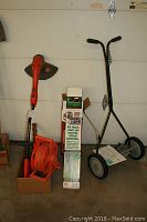 Full lot view showing GrassHog weed eater, Weed Dragon propane torch kit box, extension cord reel, and manual push mower.