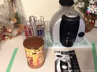 Photo of Braun Tassimo coffee maker, metal pod holder filled with coffee pods, vintage coffee tin, and some flowers in background.