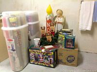Photo showing two large Rubbermaid plastic lidded bins packed with assorted gift wrapping paper rolls alongside boxed holiday decorations including a musical snow globe and outdoor light-up angel.
