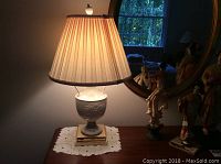 Front angle view of Lenox table lamp on wooden table with mirror and figurines in background. Lamp base is white ceramic with embossed floral detail; ivory pleated shade is turned on.