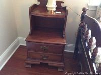 Angle view of one Pennsylvania House wooden side table showing front legs and two drawers with brass handles, upper shelf and curved back panel.