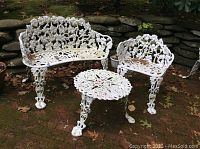 Photo showing white cast iron loveseat, chair, and round table with floral and grapevine designs, on brick patio near stone wall, with leaves scattered around