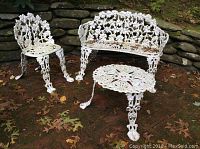 Photo showing full view of white cast iron three-piece patio set including chair, bench, and round table on brick patio with stone wall background.