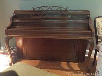 Full front view of upright piano showing honey wood finish, carved legs, and closed lid