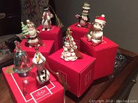 Seven Lenox Christmas ornaments arranged on and near their red Lenox boxes on a wooden surface.
