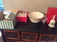 Wide shot showing all items: Lenox holiday bell, serving bowl, covered dish with decorative box, and music box candle holder on a black wooden furniture piece.