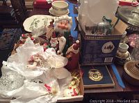 Wide shot of assorted Christmas ornaments including Santa figurines, snowman, white wrapped ornaments, and other holiday decorations spread on table with boxes and other containers.