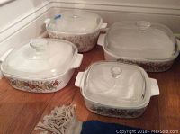 Four vintage Corningware baking dishes with fruit and vegetable decals and clear glass lids arranged on wooden floor.
