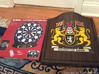 Photo showing the boxed totes electronic dartboard set next to the closed wood dartboard cabinet with Knight of Arms graphic on doors.