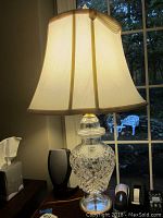 Front view of the Waterford crystal table lamp with cream shade on a table next to a window, showing crystal base patterns and gold-trimmed shade.