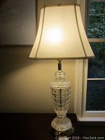Cut glass base table lamp turned on, showing illuminated white fabric shade and detailed cut glass base with brass accents.