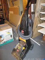 Black upright Bissell ProHeat Clearview carpet cleaner with hose attached and water tank, showing front and side views in basement setting.