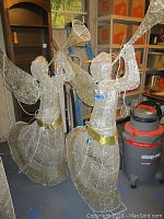 Photo showing two lighted angel yard ornaments with trumpets and gold waist accents placed indoors near shelving and a shop vacuum