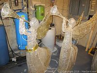 Pair of large wireframe Christmas angel figures illuminated with lights, holding trumpets, gold wings and belts, standing indoors.