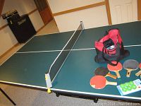 Full view of the ping pong table showing surface, net, paddles, balls, and bag with accessories.