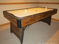 Angled view of air hockey table showing wood grain sides, black corners, and playing surface with two pushers and one puck.