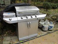 Front view of the stainless steel gas grill showing five control knobs, two doors below, side burner on left, and side shelf on right with rust spots and wear visible on the surface.
