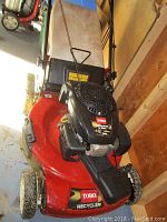 Front and side view of Toro Recycler lawnmower showing red deck, black engine cover with Toro branding, and beige grass collection bag.
