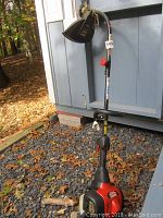 Full view of Craftsman weedwacker leaning against blue shed on gravel with some dried leaves visible around it.